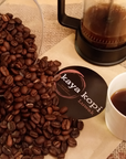 Coffee beans, a French press, and a cup of coffee on a table with a 'kaya kopi' coaster.