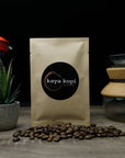 Kaya Kopi coffee bag with coffee beans and coffee-making equipment on a wooden surface.