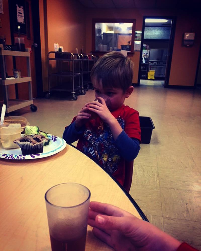 Child sitting at a table with a plate of food and a glass in a casual setting.