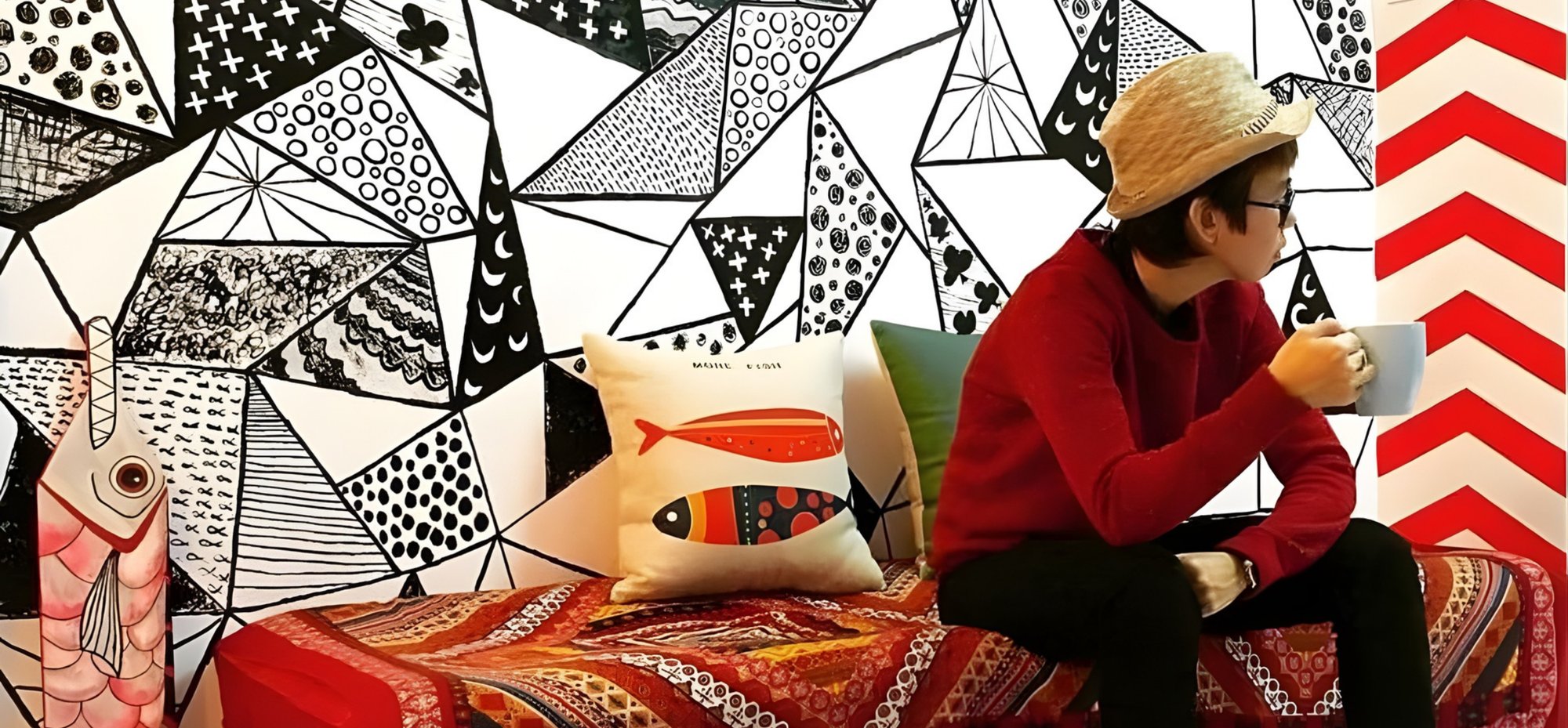 Person sitting on a bed with colorful bedding and pillows against a geometric patterned wall and enjoying a cup of luwak coffee.